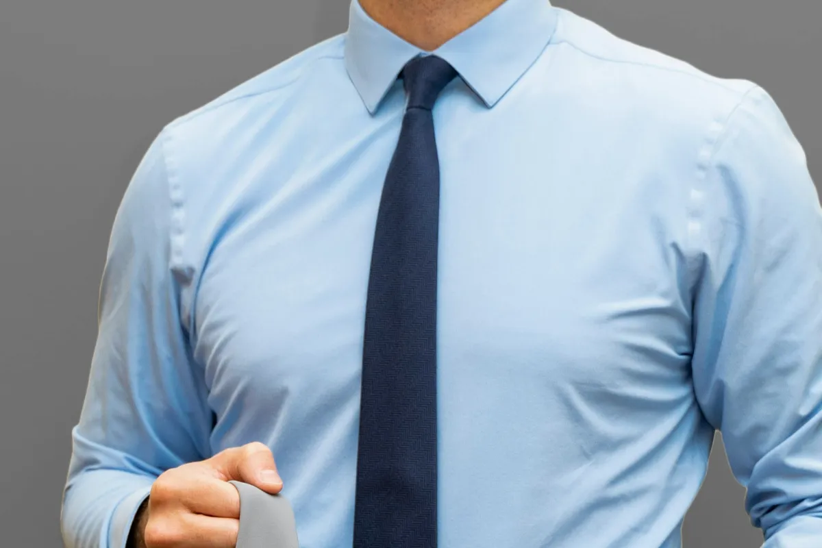 Man in a light blue business shirt and navy tie, holding a grey suit jacket over his arm
